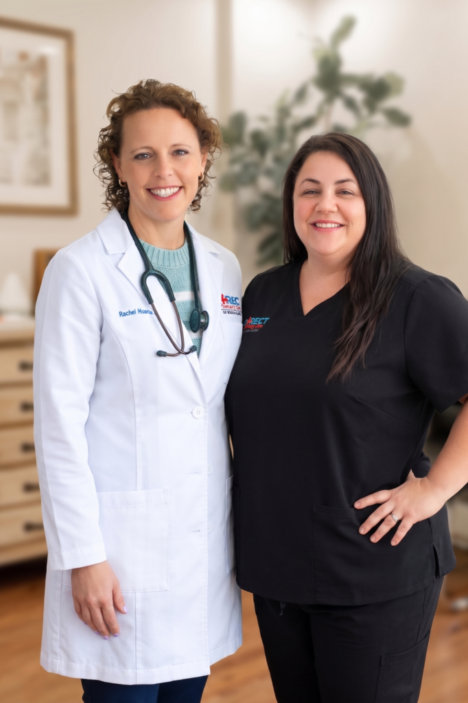 Two healthcare professionals stand side by side in a medical office, smiling at the camera while wearing branded clinical attire.