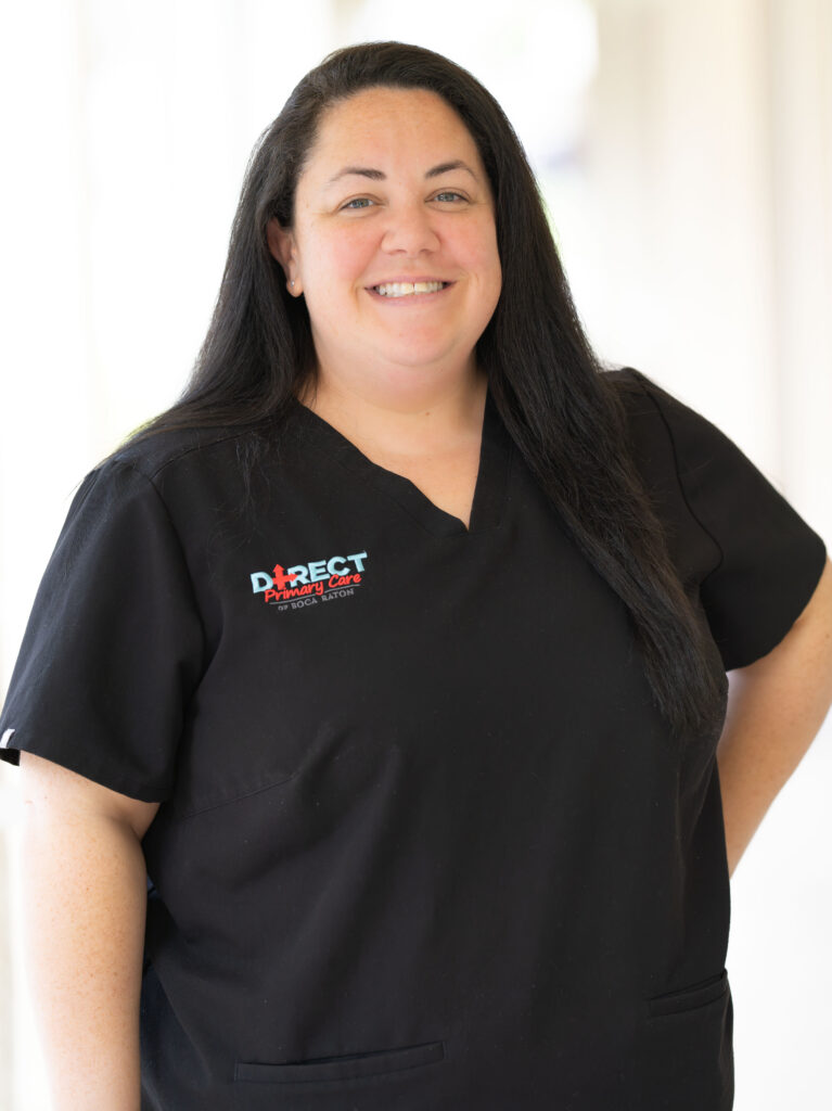 Smiling woman with long dark hair wearing black Direct Primary Care of Boca Raton scrubs in a bright office.